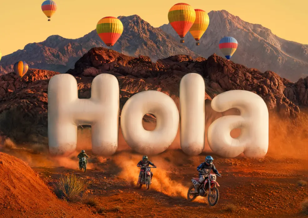 Ciclistas de motocross subiendo por un sendero en el desierto, alejándose de unas letras gigantes inflables que forman la palabra "¡Hola!", mientras globos aerostáticos de colores flotan sobre montañas rocosas.